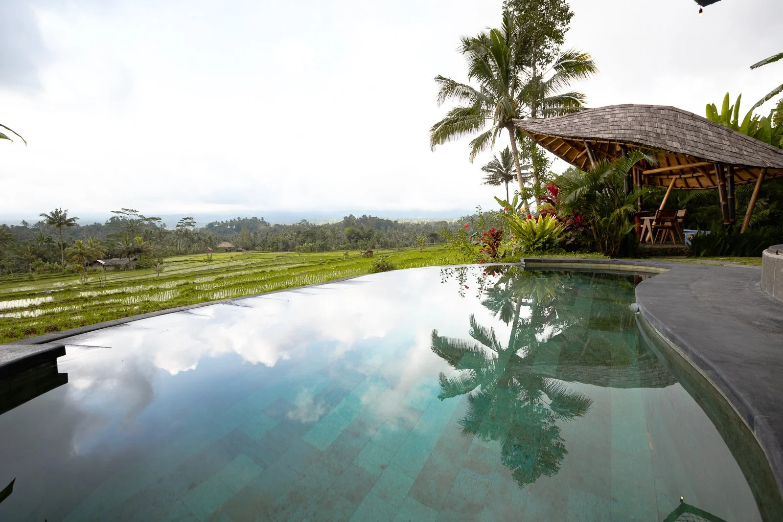 Piscine à débordement avec vue sur des champs de riz et une cabane en bambou entourée de végétation tropicale, sous un ciel nuageux.