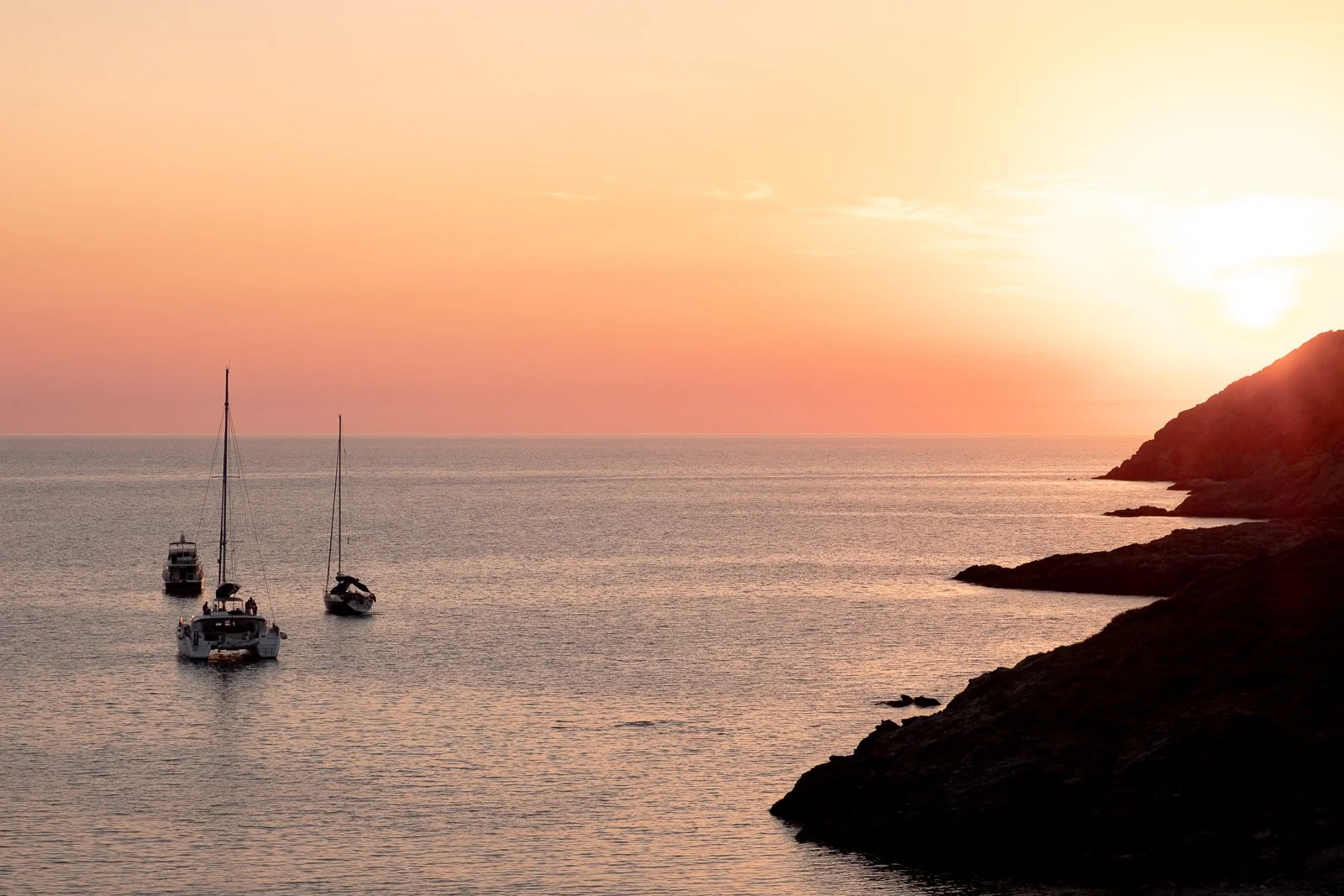 Coucher de soleil sur la mer avec trois bateaux et une côte rocheuse.