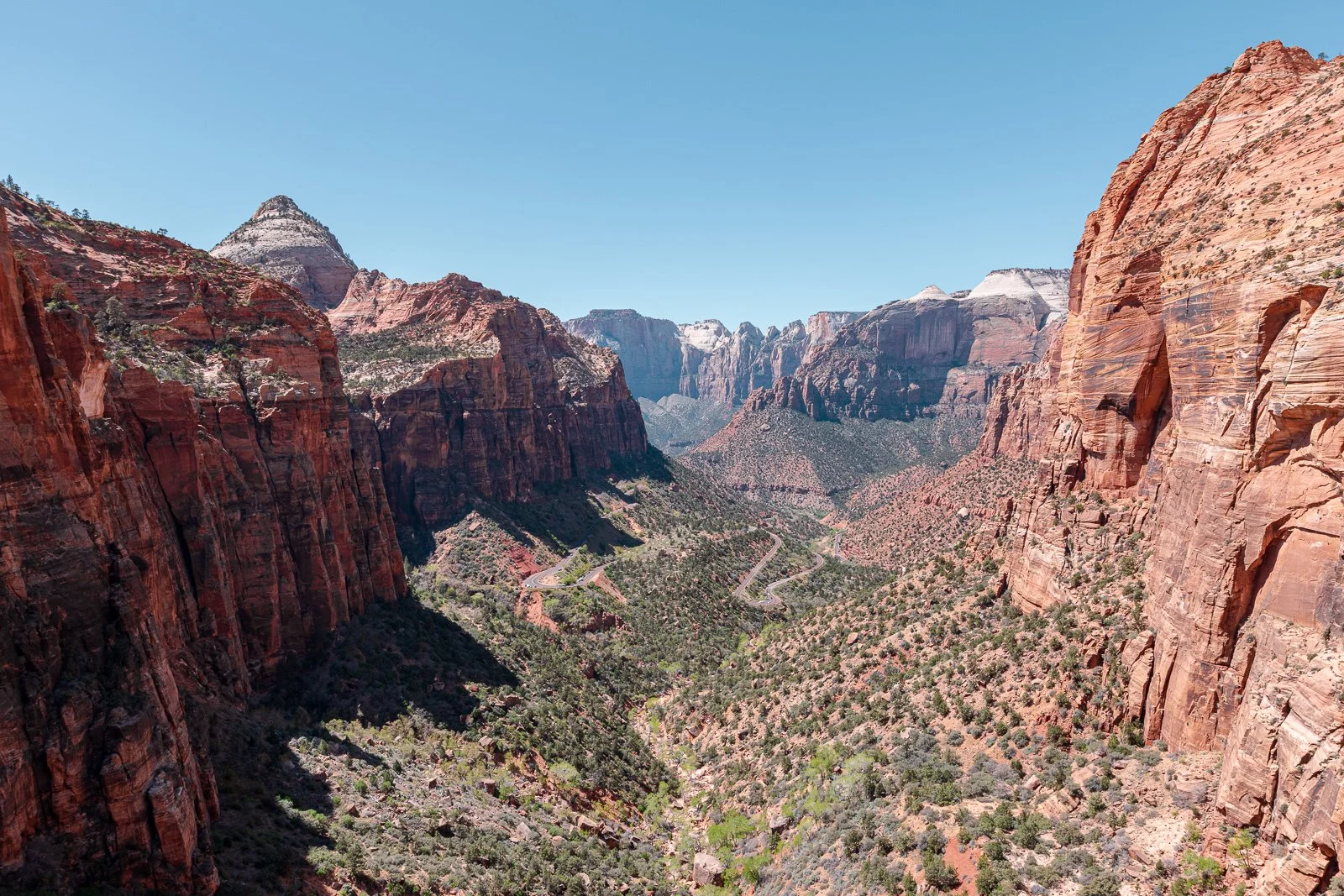 Canyon avec parois rocheuses rouges et végétation sparse, route serpentant en bas, ciel bleu clair.