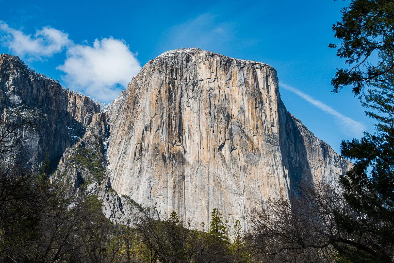 Une montagne imposante avec une surface rocheuse vertigineuse, encadrée par des arbres et un ciel bleu avec quelques nuages.