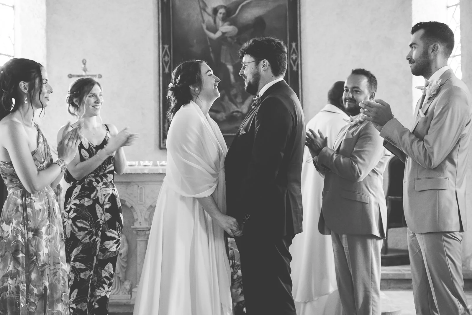 Un mariage avec un couple échangeant des vœux devant un prêtre, entouré d'amis qui applaudissent dans une église.