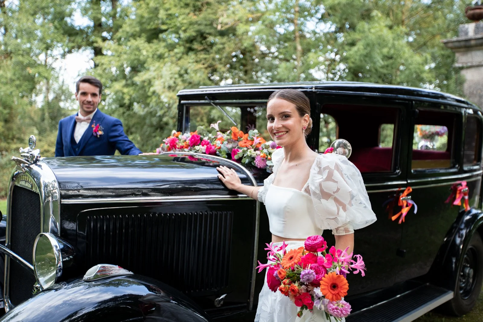 Un couple de mariés, la femme en robe blanche et l'homme en costume bleu, pose à côté d'une voiture ancienne décorée de fleurs colorées dans un environnement verdoyant.