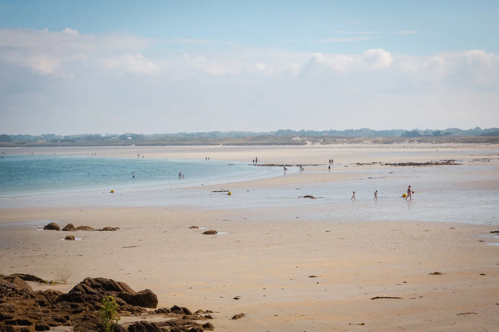 Plage avec sable, eaux calmes, personnes marchant et nageant dans la mer, ciel partiellement nuageux.