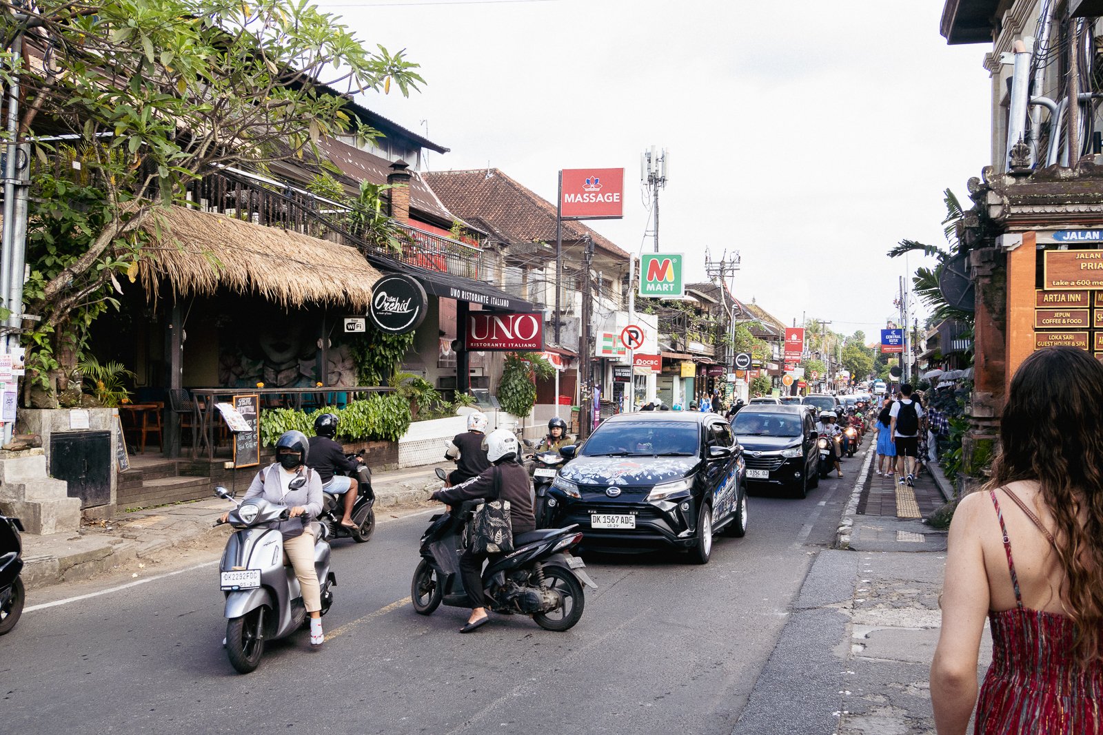 Une rue animée avec des véhicules et des motos, des commerces en arrière-plan, et des piétons marchant sur le trottoir. L'ambiance suggère une ville touristique en Indonésie.
