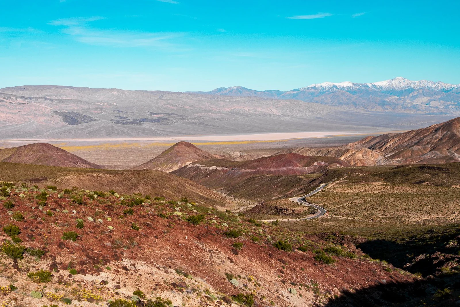 Paysage désertique avec des montagnes colorées, une route sinueuse et des montagnes enneigées à l'arrière-plan, sous un ciel bleu clair.