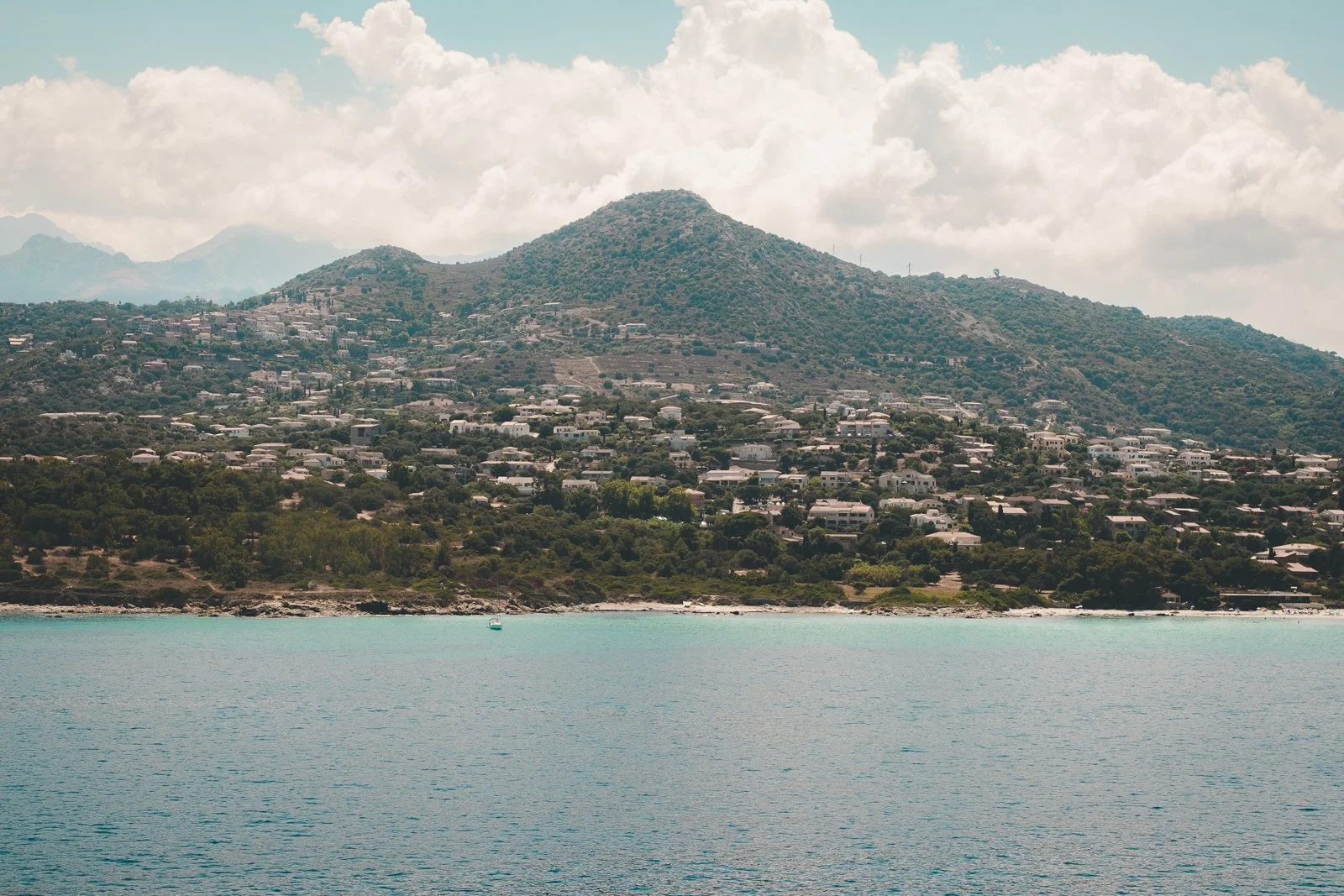 Une montagne avec des maisons en contrebas près d'un lac ou d'une mer, avec un ciel partiellement nuageux.