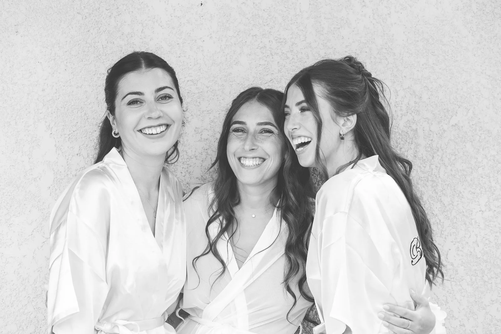 Trois femmes souriantes et riant, portant des robes satinées, posant contre un mur texturé.