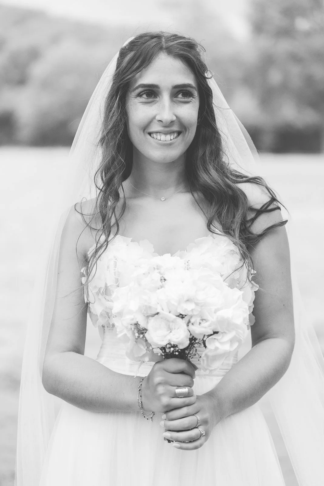 Une femme en robe de mariée tenant un bouquet de fleurs, souriant, avec un voile derrière elle, en extérieur.