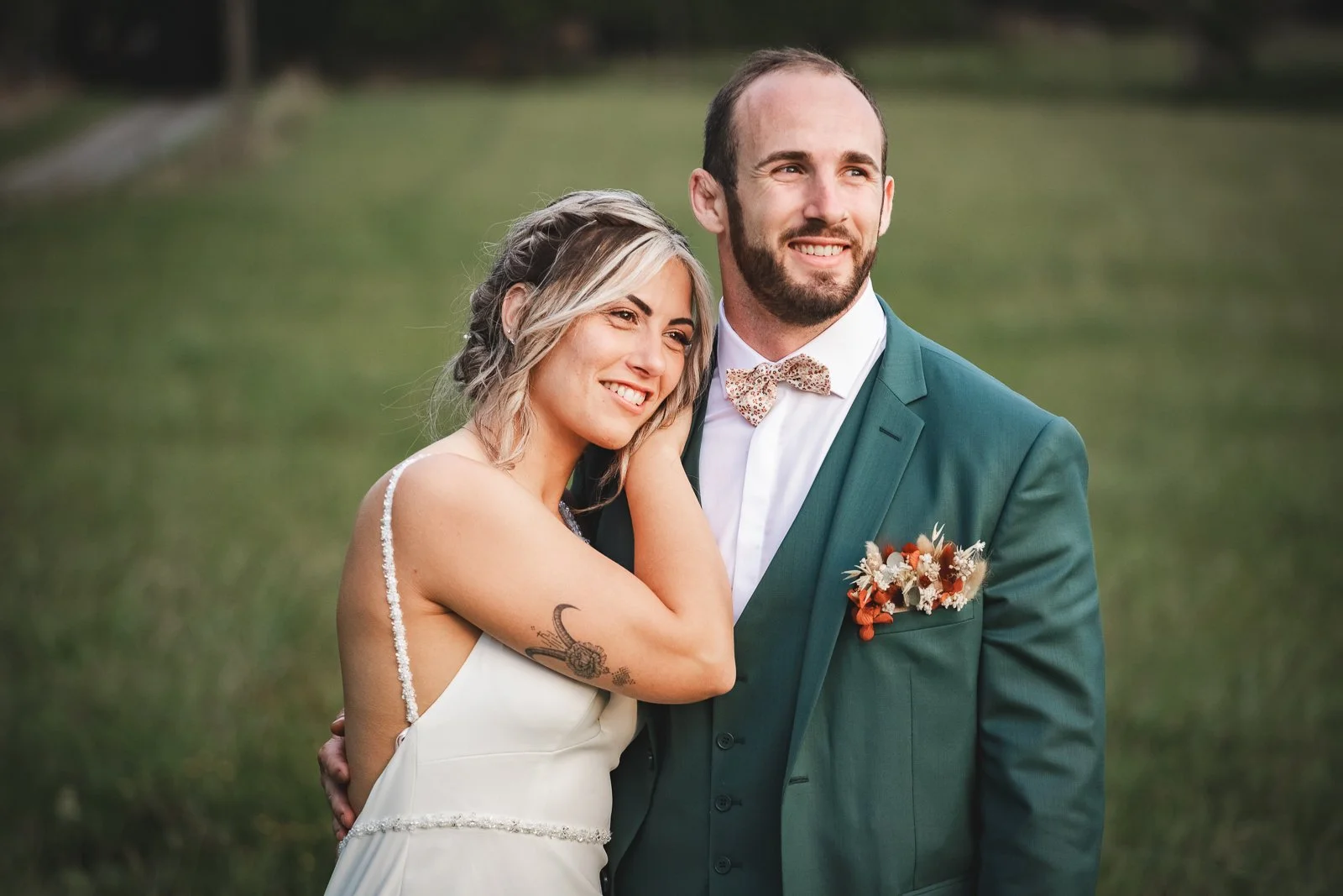 Un couple de mariage pose en plein air, la femme avec une robe blanche et un tatouage sur le bras, l'homme en costume vert avec un nœud papillon à motifs, entourés d'un paysage verdoyant.
