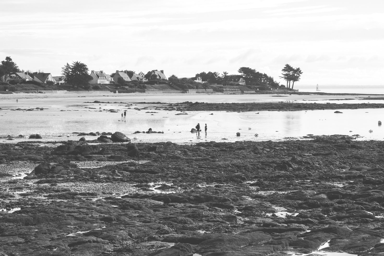 Plage rocheuse avec des personnes marchant et une maison en arrière-plan, en noir et blanc.