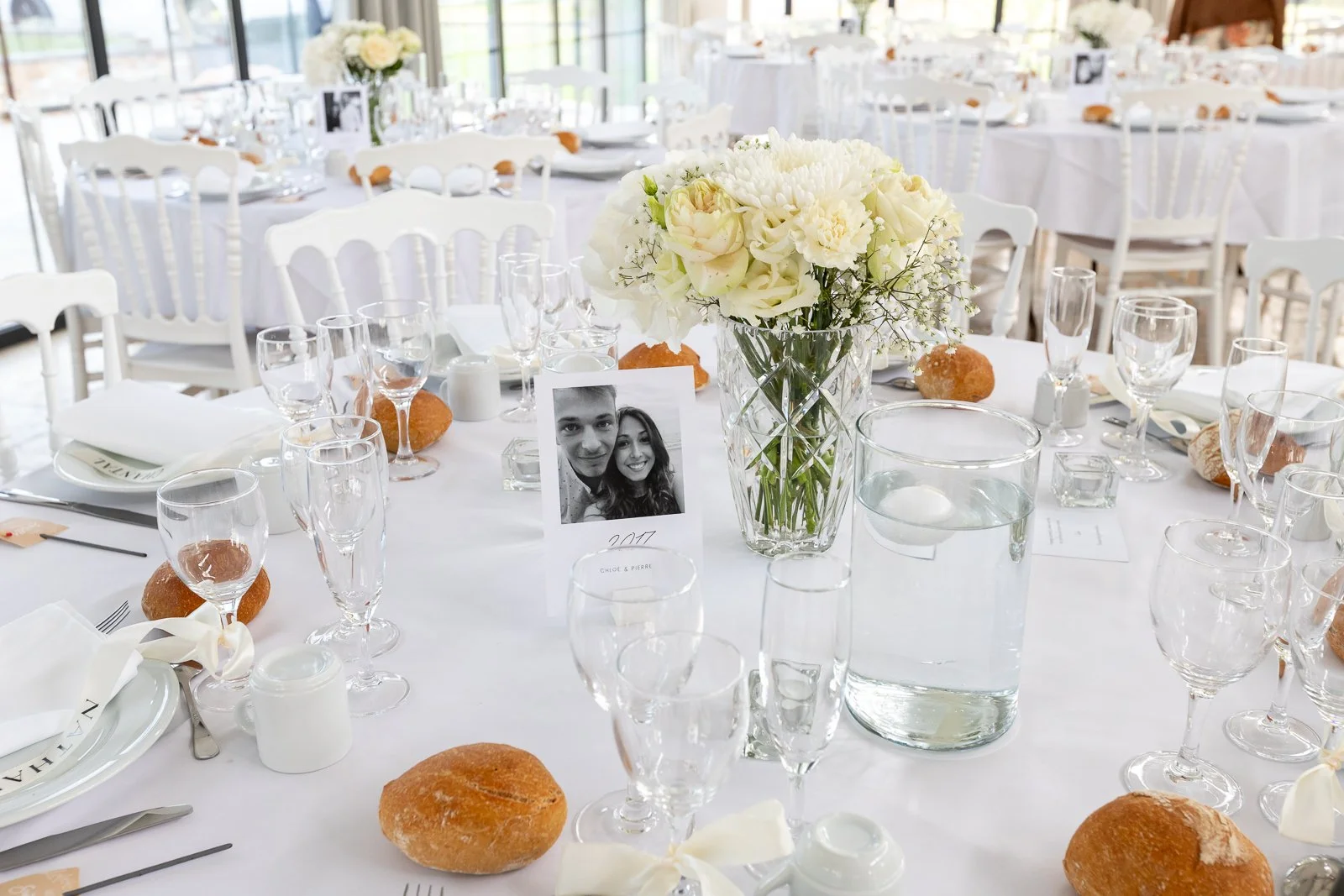 Table de banquet décorée avec un vase de fleurs blanches, des assiettes, des verres à vin et à eau, des petits pains, une photo de couple, et une place de fête, dans une ambiance lumineuse.