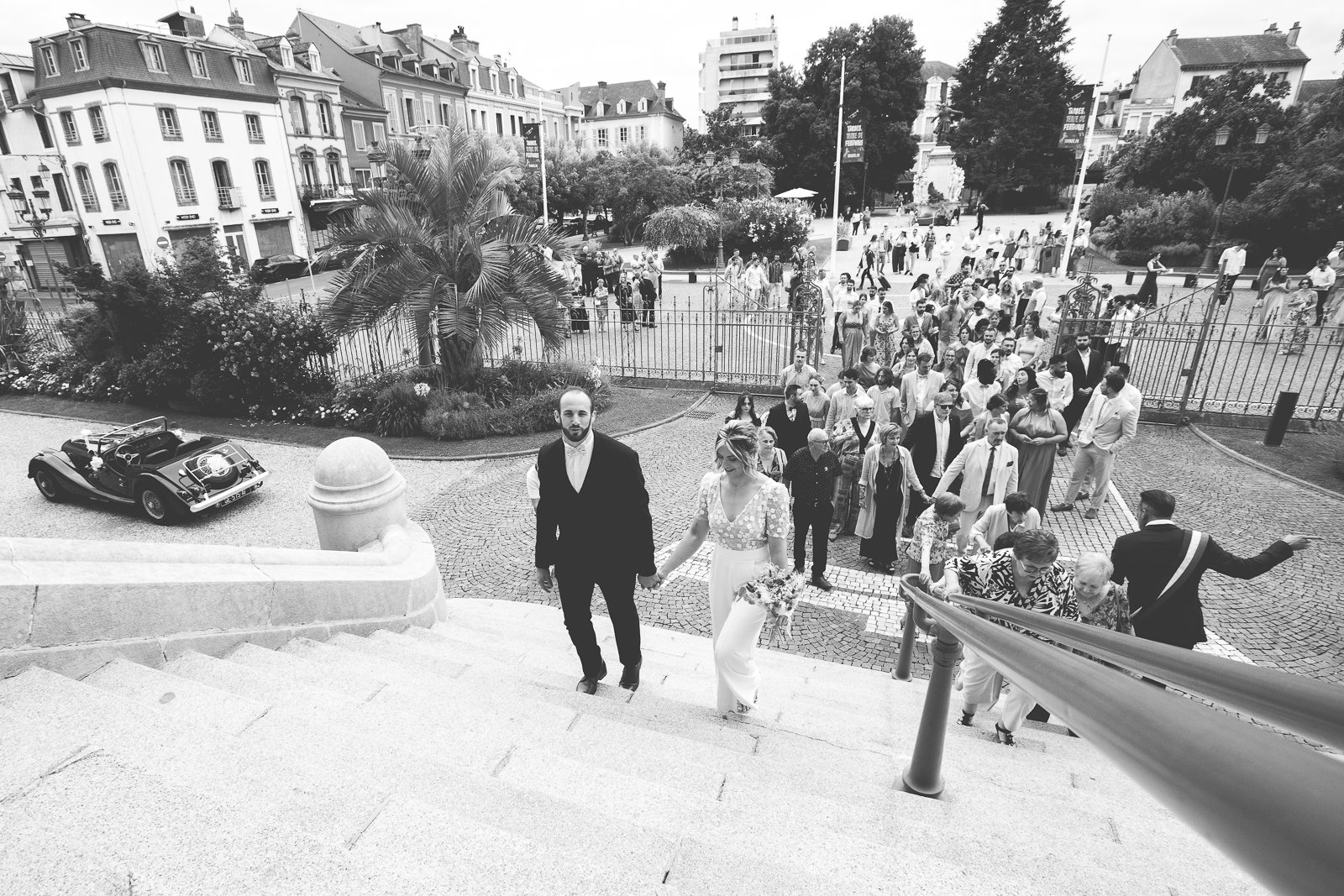 Mariage avec le couple principal montant les escaliers, entouré d'invités, dans une ville avec bâtiments anciens et une voiture de collection garée à côté.