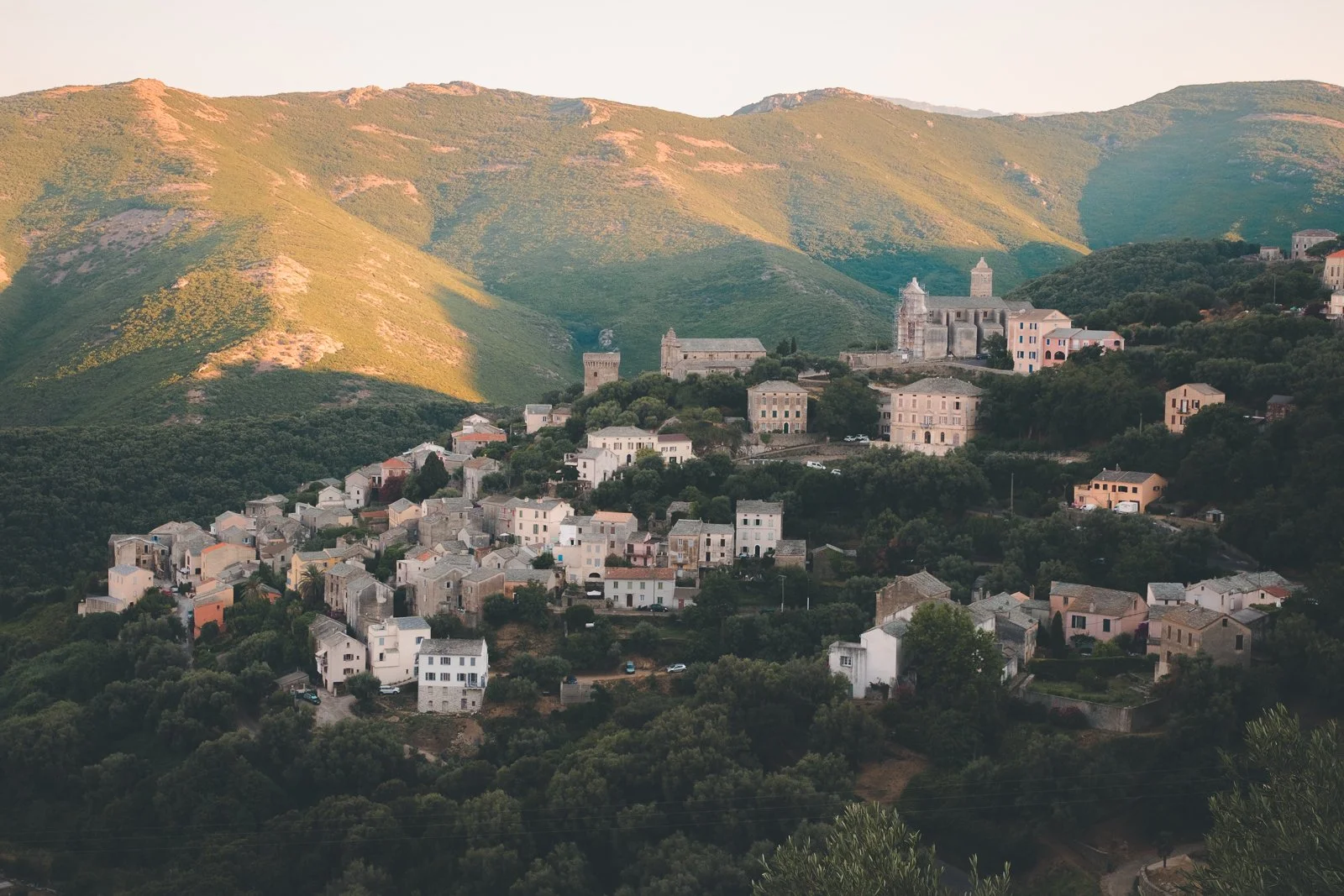 Petit village de montagne avec des maisons en pierre ou en béton, entouré de verdure et surplombant une vallée, avec un château ou une forteresse ancienne sur la colline au fond.