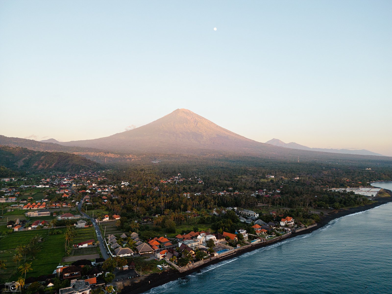 Vue aérienne d'une ville côtière avec un volcan en toile de fond, le ciel clair et une mer calme.