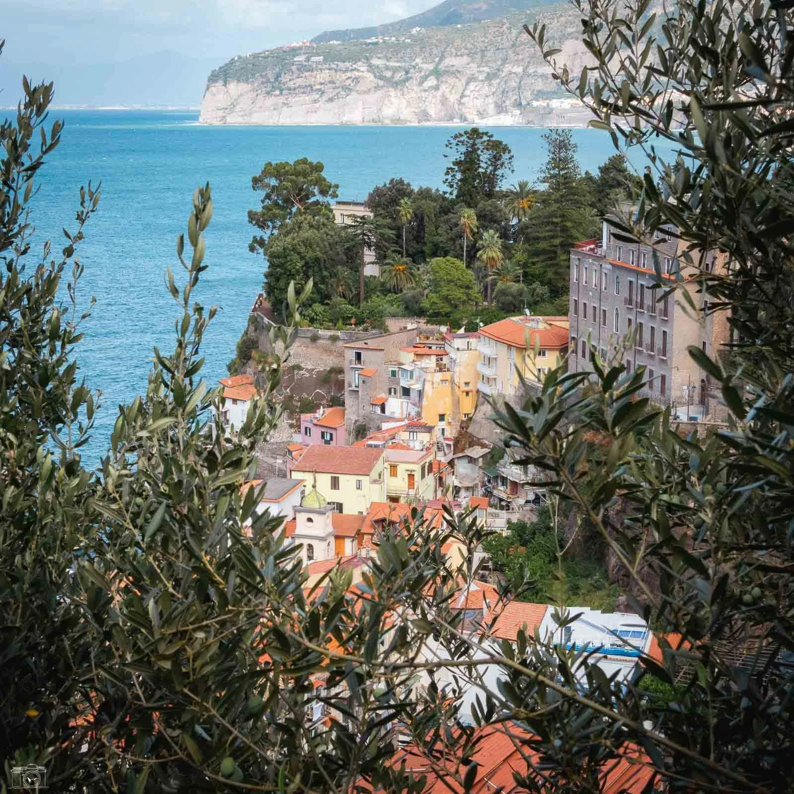 Village côtier avec maisons aux toits rouges, entouré de végétation abondante, et vue sur la mer avec une falaise en arrière-plan.