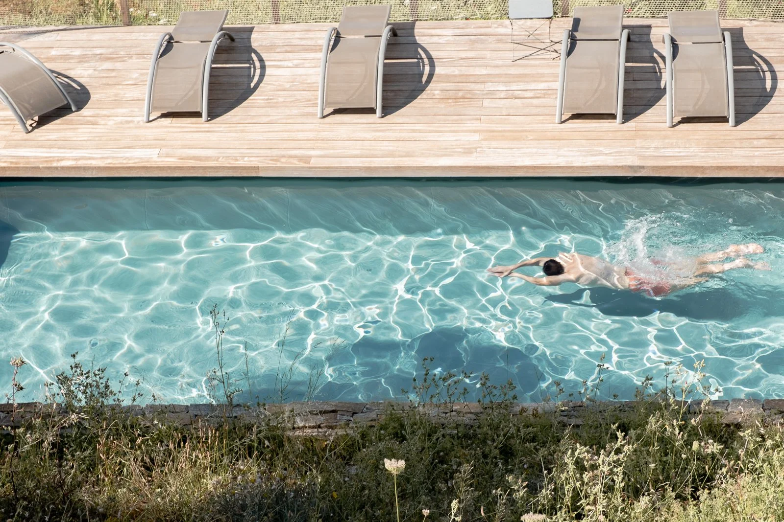 Un homme nage dans une piscine turquoise claire, bordée de planches en bois et quelques chaises longues en arrière-plan.