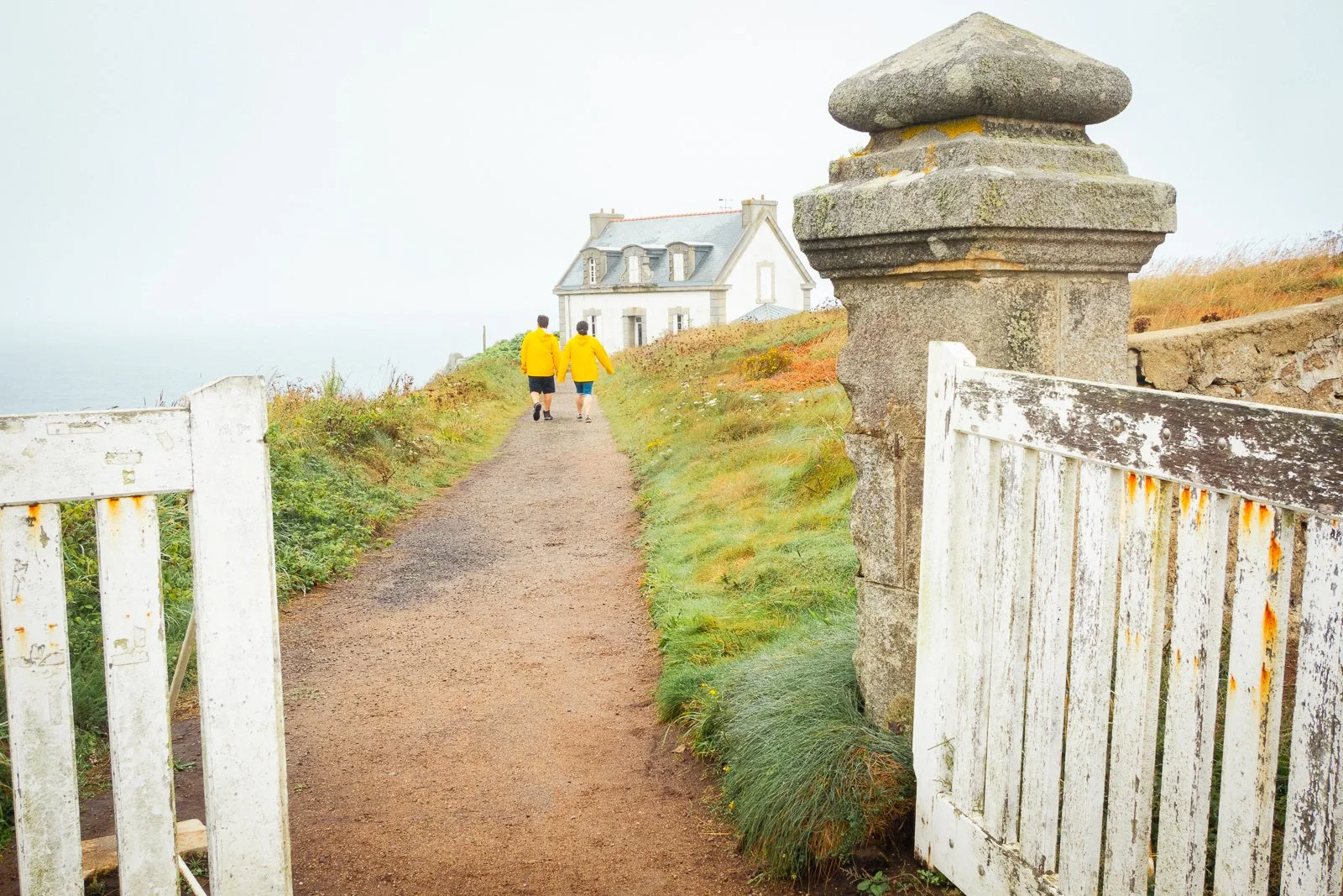 Deux personnes en jaune marchent vers une maison blanche au bord de la mer, à travers un chemin de terre, avec une vieille clôture en bois blanc et un pilier en pierre sur le côté droit.