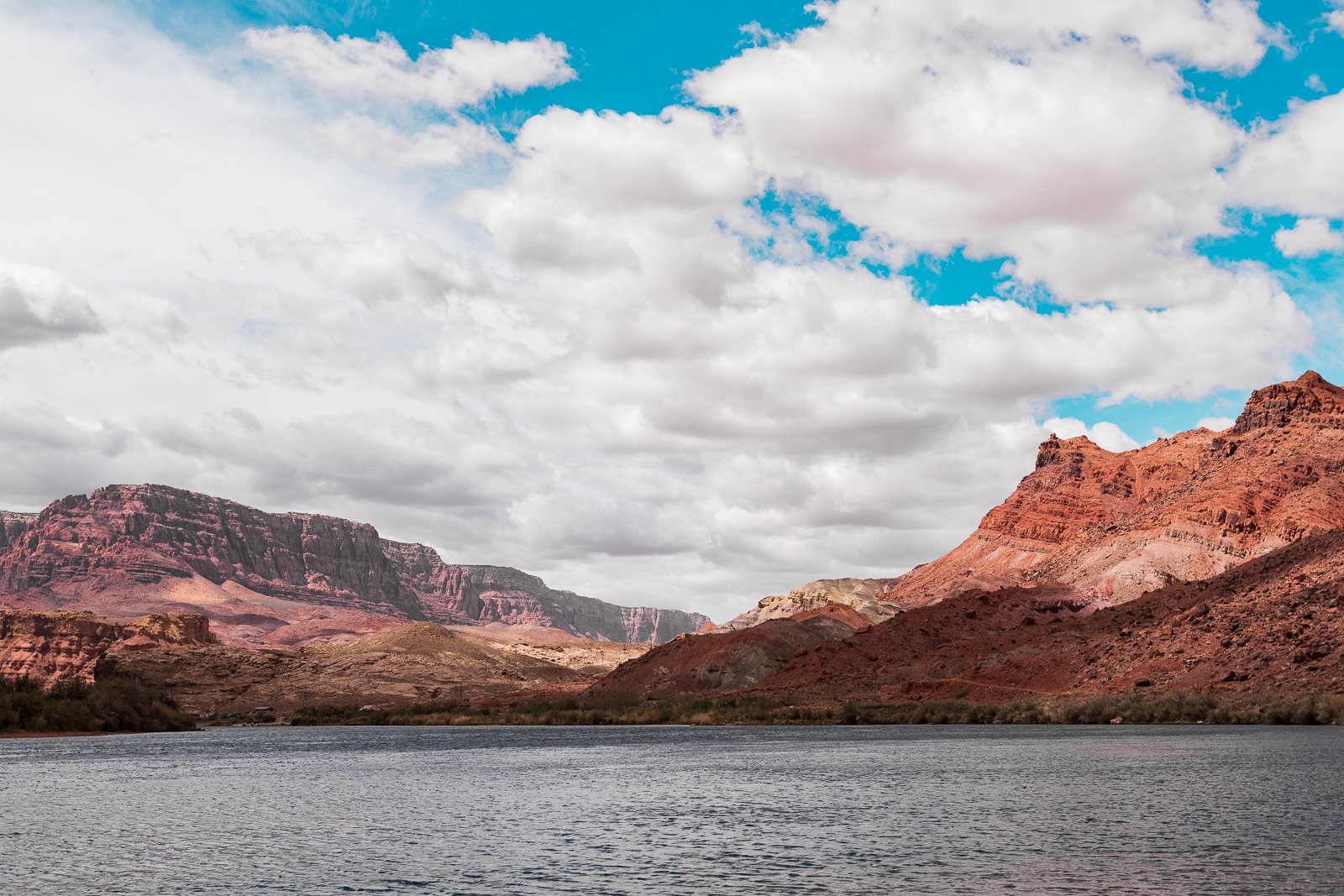 Paysage avec un lac au premier plan, montagnes rouges et brunes en arrière-plan, ciel partiellement couvert de nuages blancs et quelques nuages bleus