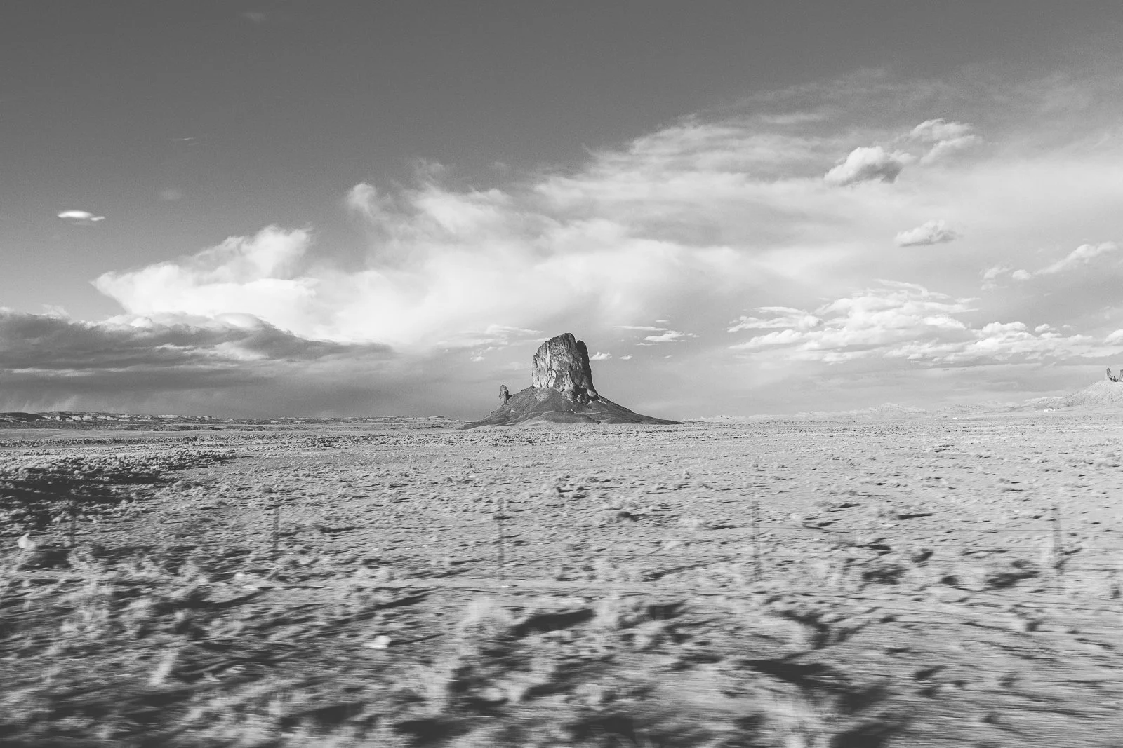 Paysage désertique avec une formation rocheuse distinctive au centre, ciel nuageux, en noir et blanc.