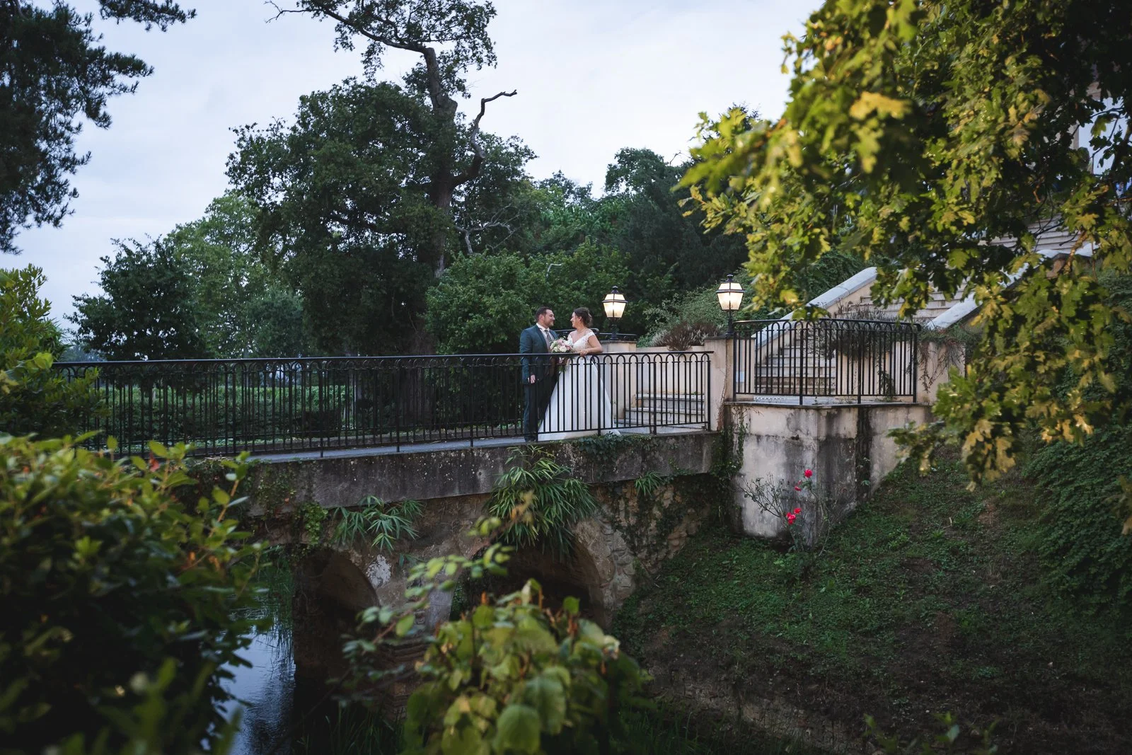 Un couple de mariés sur un pont en pierre surplombant une petite rivière, entouré d'arbres et de lampadaires, lors d'une journée nuageuse.