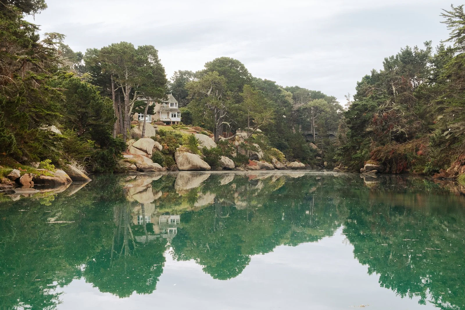 Maison blanche entourée d'arbres et de rochers, reflet dans un lac calme.