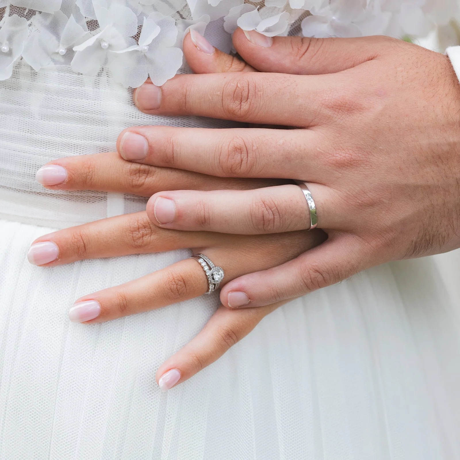 Les mains d'un nouveau couple de mariés portant des alliances, posées l'une sur l'autre avec une robe blanche en arrière-plan.