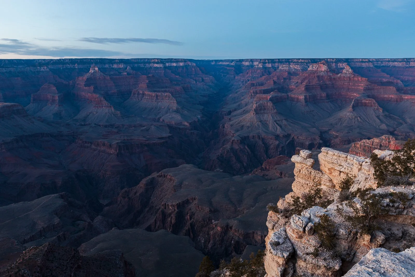 Vue du Grand Canyon au crépuscule avec un ciel clair et quelques nuages, montrant les formations rocheuses colorées et profondes du canyon.