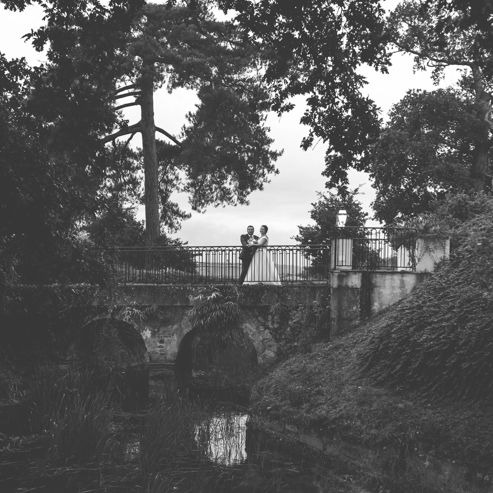 Un couple de mariés en costume et robe de mariée, debout sur un pont en fer forgé au-dessus d'une rivière, entouré d'arbres et de verdure, dans un cadre naturel.