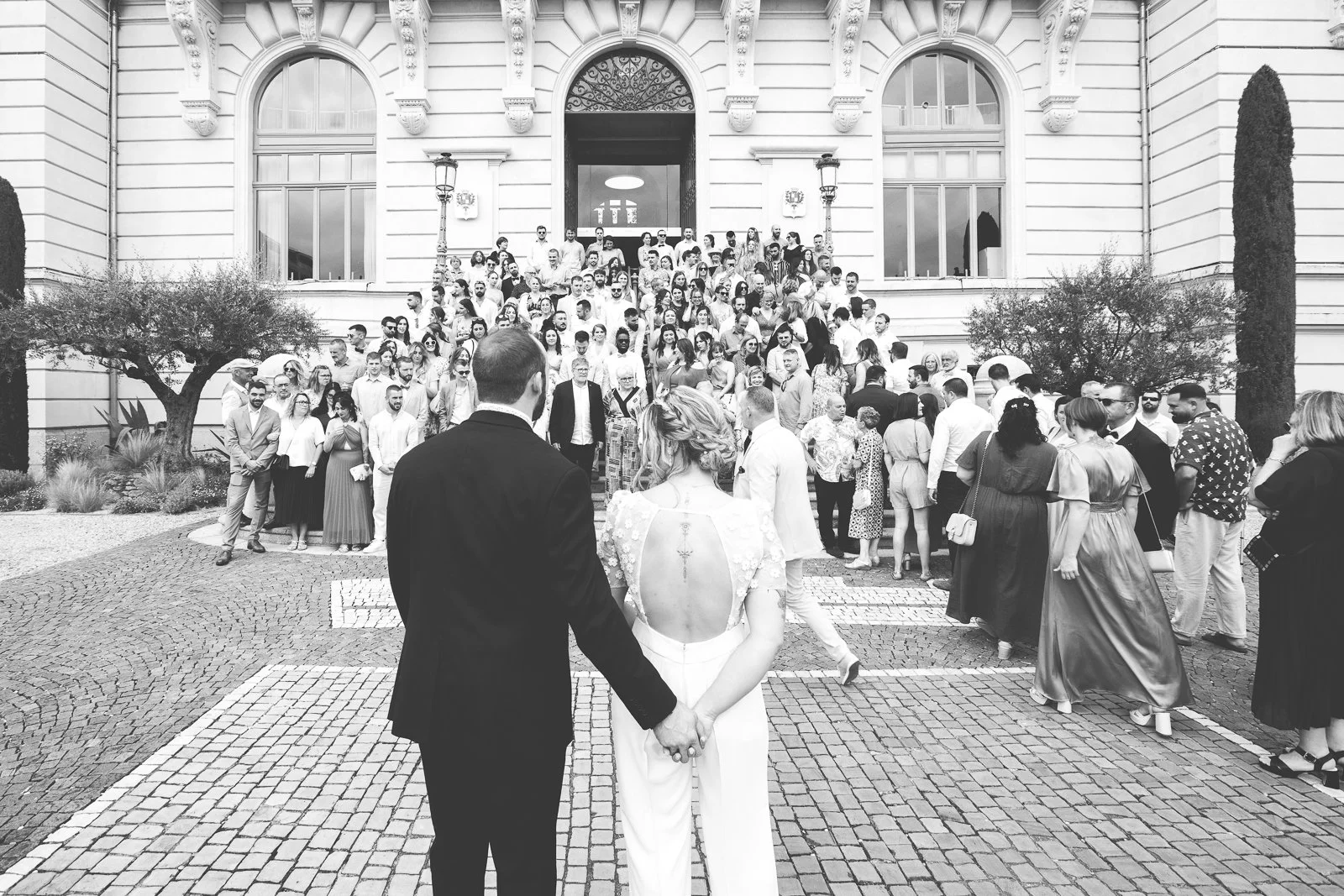 Un mariage avec un couple en première plan, devant une grande foule rassemblée devant un bâtiment officiel.