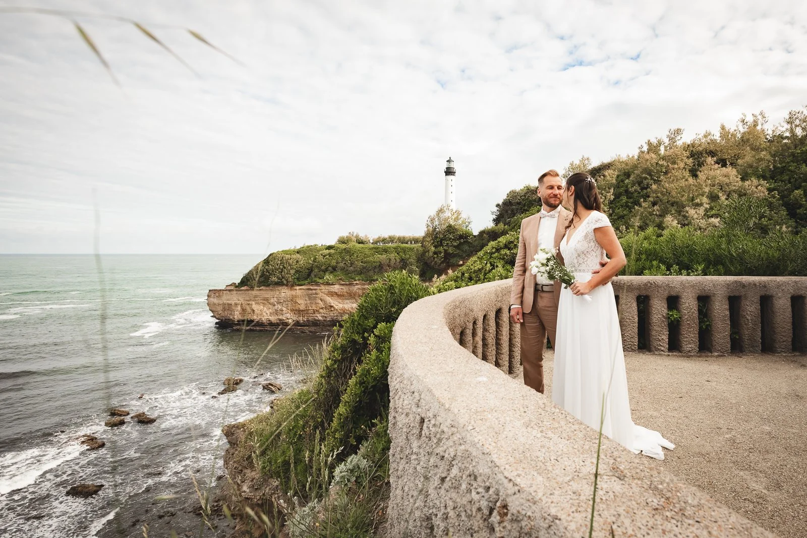 Un couple en mariage se tient sur une promenade en bord de mer, avec un phare sur une colline en arrière-plan, entourés de verdure et d'une mer calme.