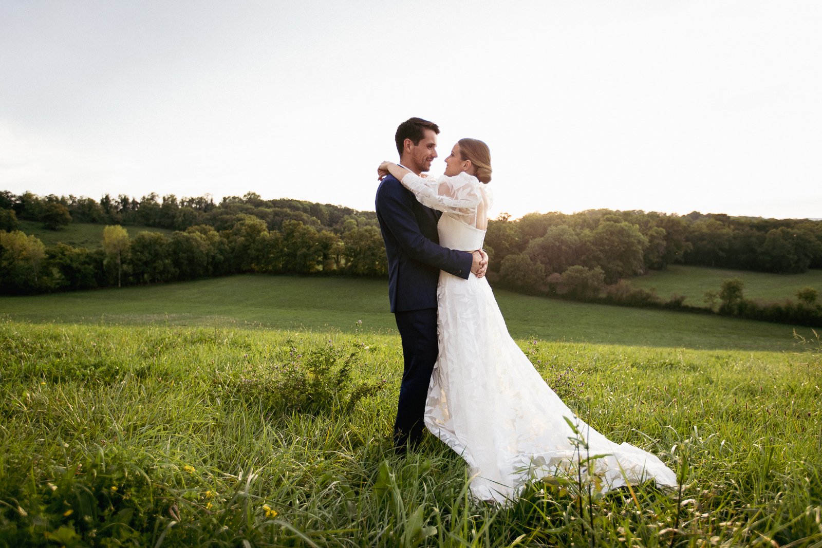 Un couple en costume de mariage se tient dans un champ vert, s'embrassant tendrement, avec une forêt en arrière-plan et un ciel clair au crépuscule.