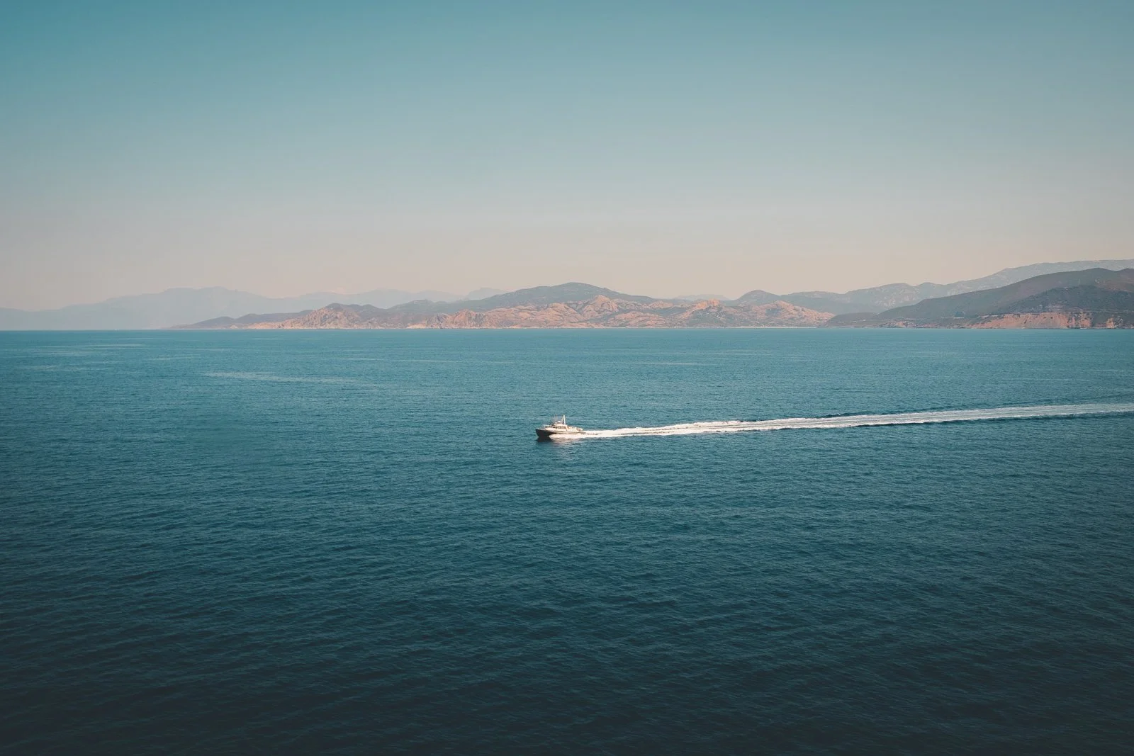 Vue de la mer avec un bateau à moteur laissant une traînée derrière lui, des montagnes au loin sous un ciel clair.