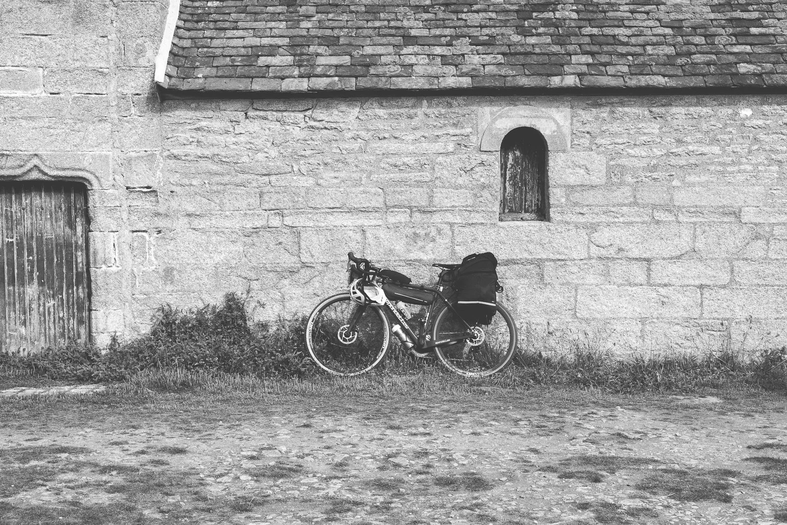 Une photo en noir et blanc d'un vélo avec sac de voyage, stationné contre un vieux mur en pierre, avec une petite fenêtre en arc et une porte en bois.