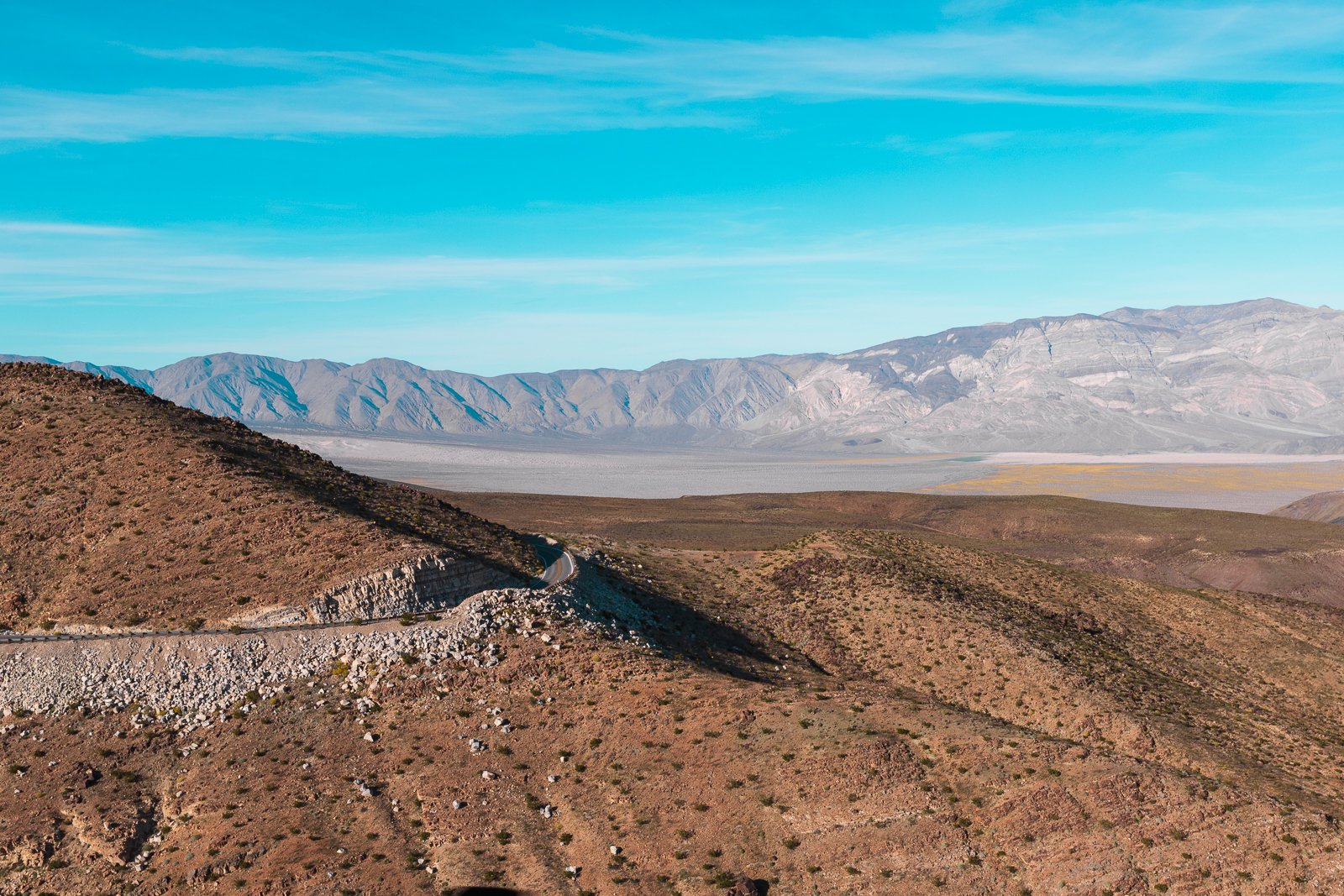 Paysage désertique avec montagnes au loin, route sinueuse serpentant à travers la terre aride, ciel bleu clair avec quelques nuages