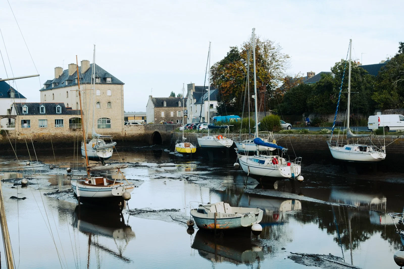 Bateaux de plaisance amarrés dans un port en maree basse, avec bâtiments anciens en arrière-plan, entouré d'arbres et de voitures.