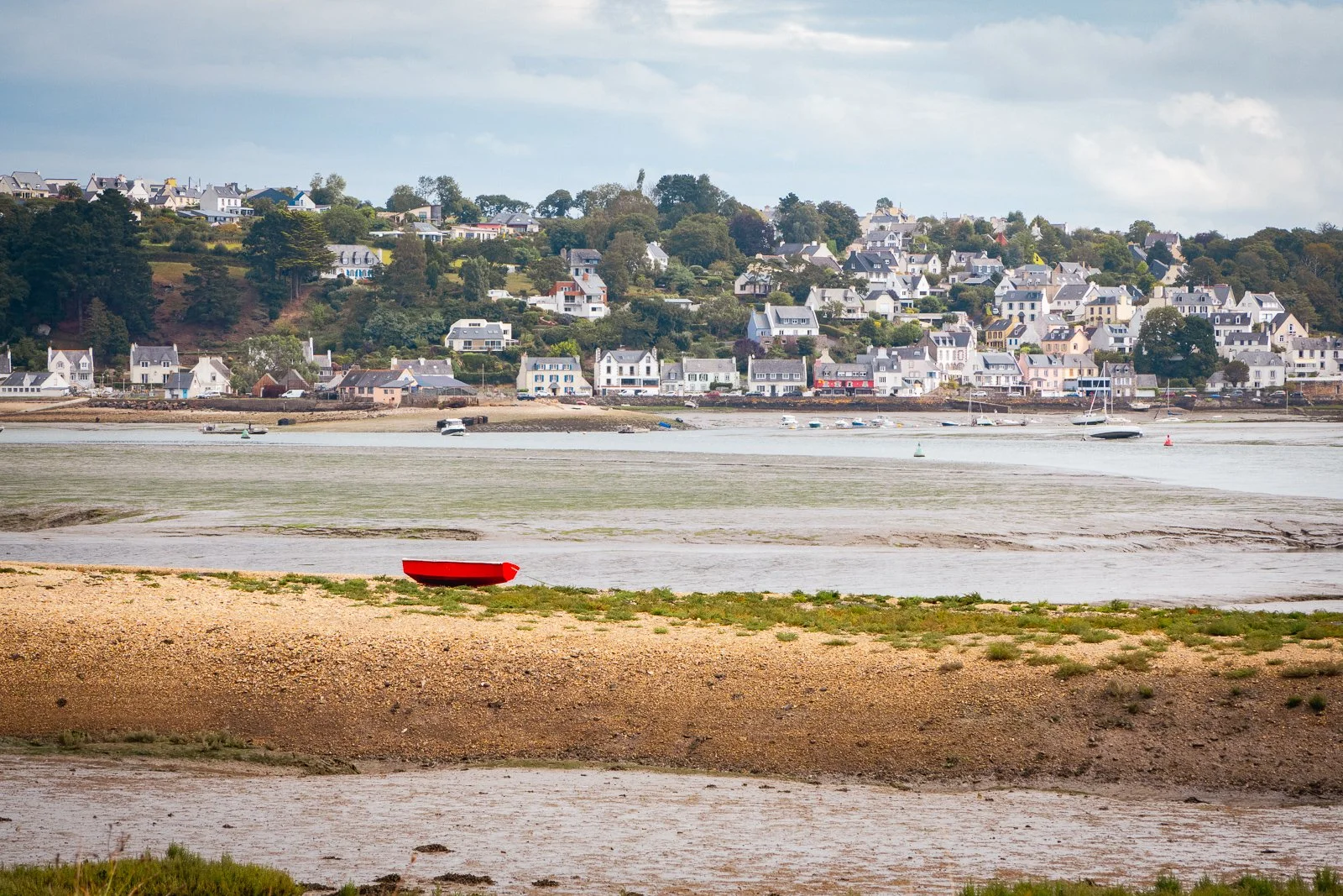 Vue d'une côte avec des maisons en arrière-plan, un bateau rouge sur la plage en premier plan, et des bateaux dans l'eau
