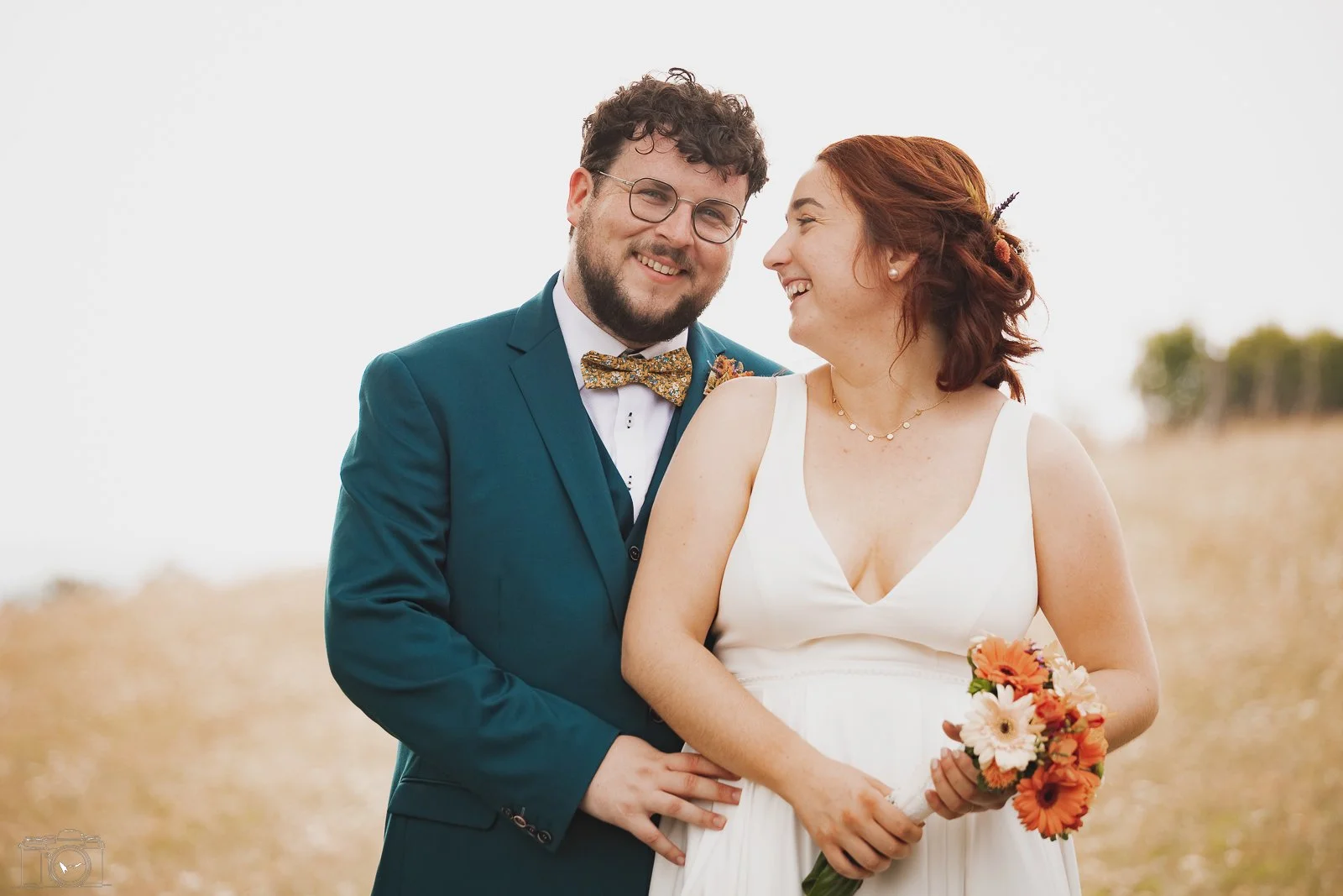 Un couple lors d'un mariage, la femme en robe blanche et le homme en costume vert avec un nœud papillon à motifs floraux, ils sourient et regardent l'un l'autre, dans un champ ouvert.
