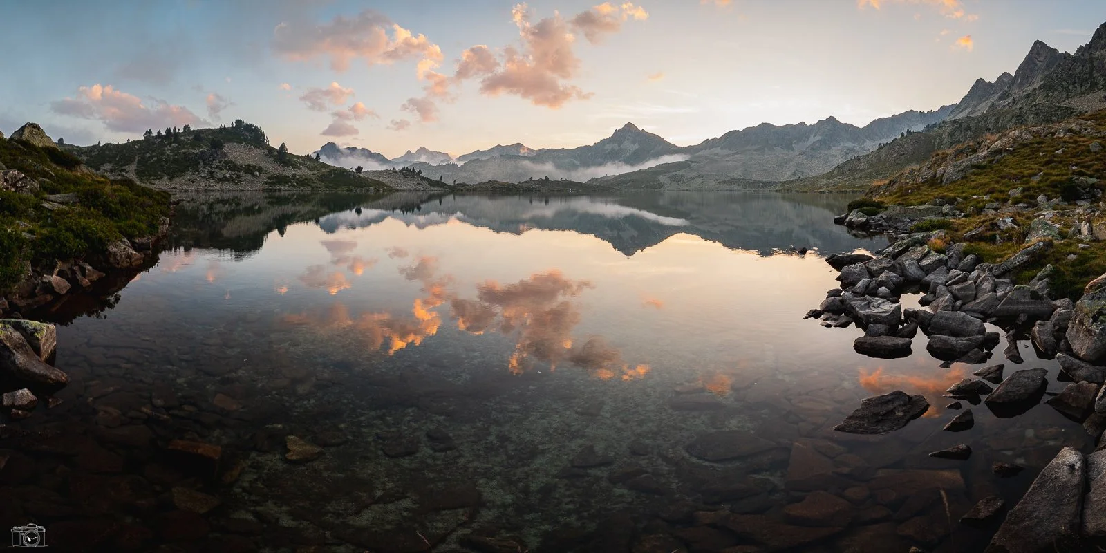 Un lac de montagne calme avec des rochers au bord, des montagnes en arrière-plan, et un ciel nuageux aux couleurs pastel, reflétant l'image dans l'eau.