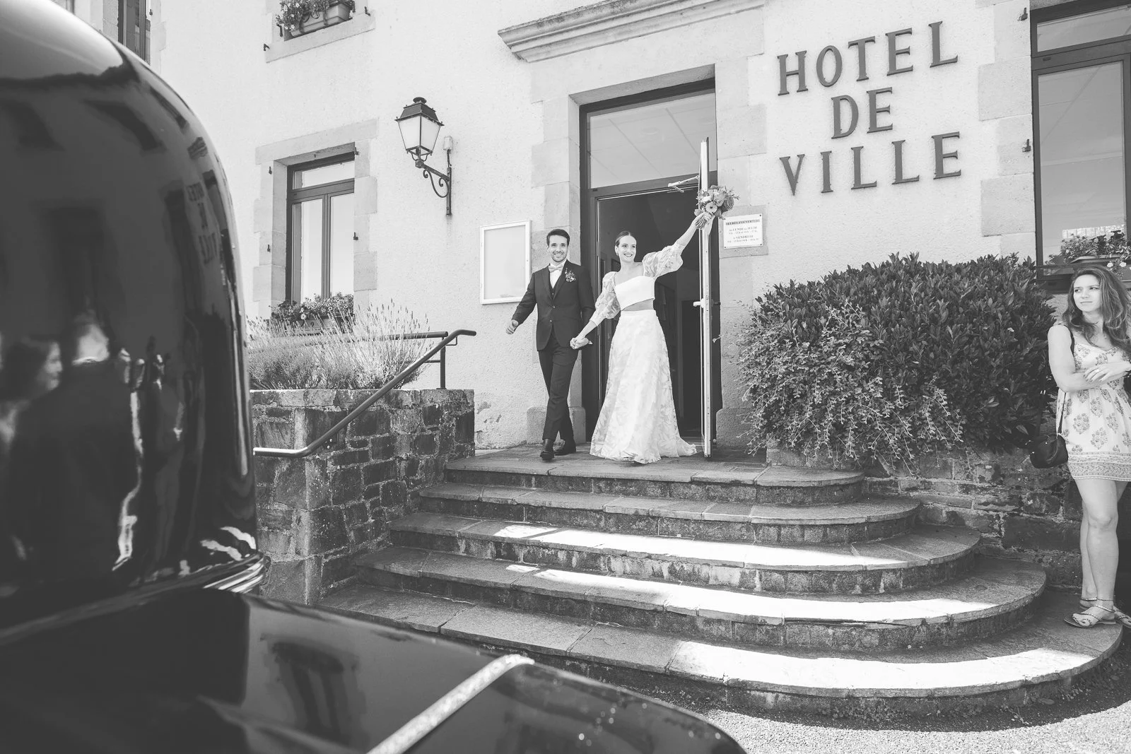 Un couple de mariés sortant de l'hôtel de ville, la mariée porte une robe blanche avec un bouquet, et l'homme porte un costume noir. Une femme se trouve à droite avec un sac à main. La scène est en noir et blanc.