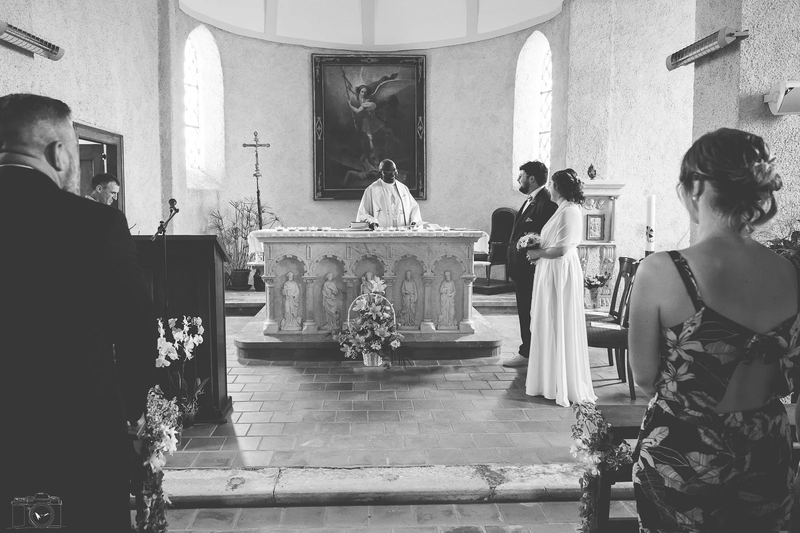 Cérémonie de mariage dans une église, couple devant le prêtre, invités observeurs, décor floral et peinture religieuse derrière l'autel