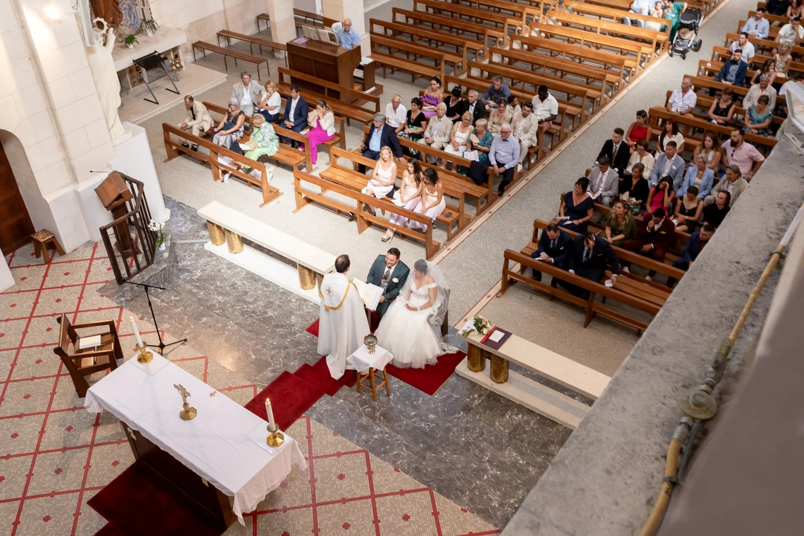Cérémonie de mariage dans une église avec l'officiant, le couple marié et des invités assis dans les bancs.