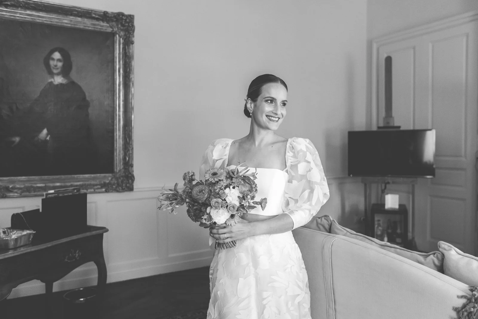 Une femme en robe de mariée tenant un bouquet de fleurs, souriante, dans une pièce élégante avec un portrait encadré au mur et un téléviseur.