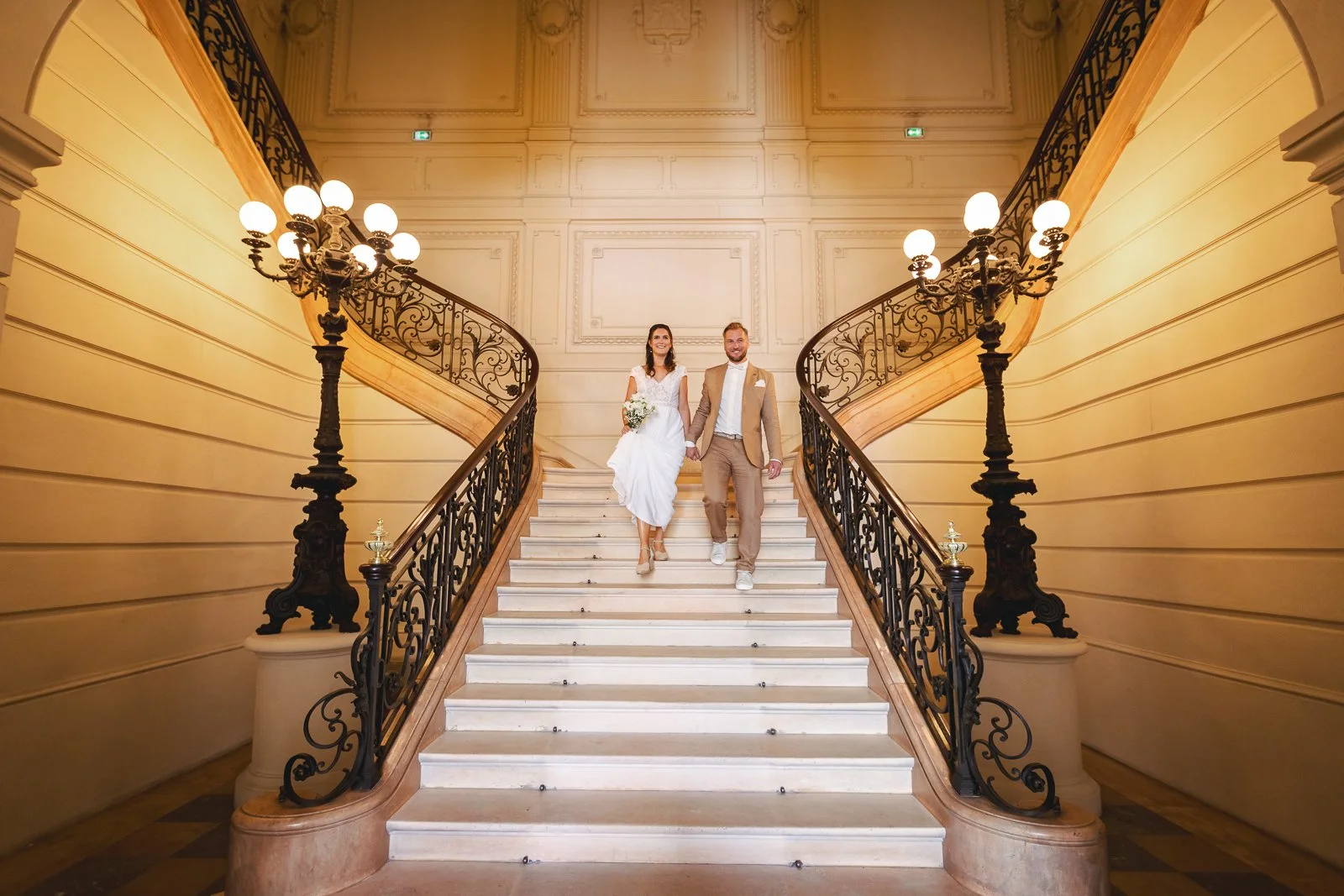 Un couple en mariage descend un escalier en marbre dans un bâtiment élégant avec des lampadaires en fer forgé.