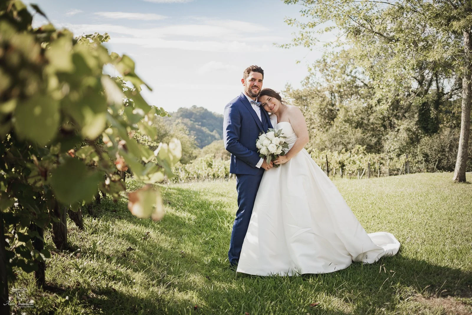 Un couple de mariés posant dans un champ verdoyant en plein air, avec un arbre à gauche et des collines en arrière-plan, le jour ensoleillé.