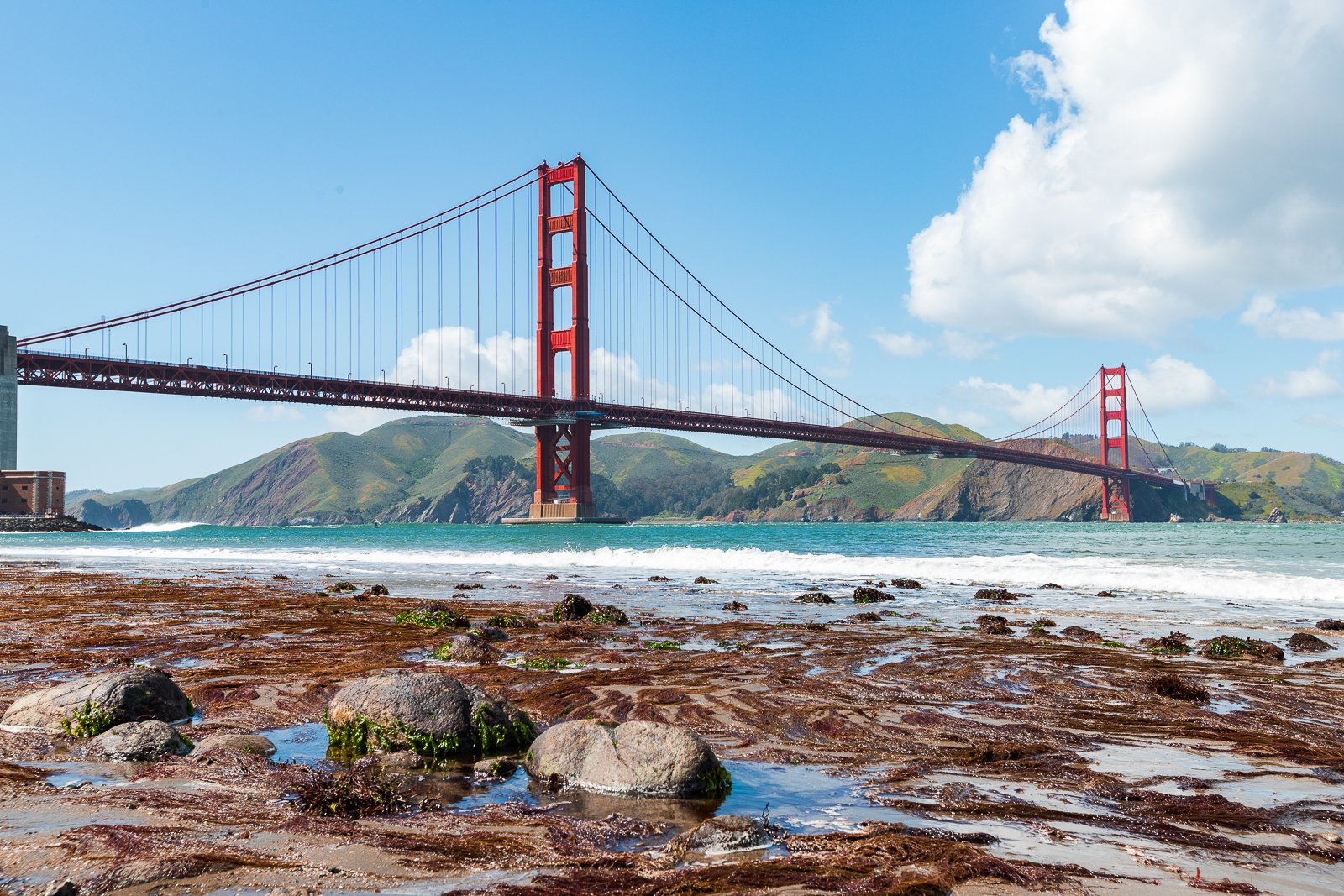 Le pont Golden Gate rouge au-dessus de l'océan avec des collines verdoyantes en arrière-plan, ciel bleu et quelques nuages.