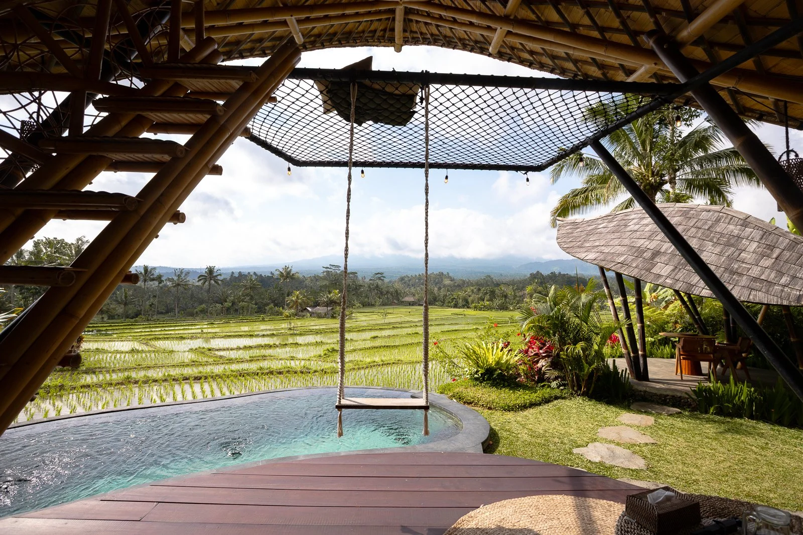 Une vue d'une terrasse en bois avec un hamac suspendu, une piscine, et un paysage de champs de riz avec des palmiers en arrière-plan, sous un ciel partiellement nuageux.