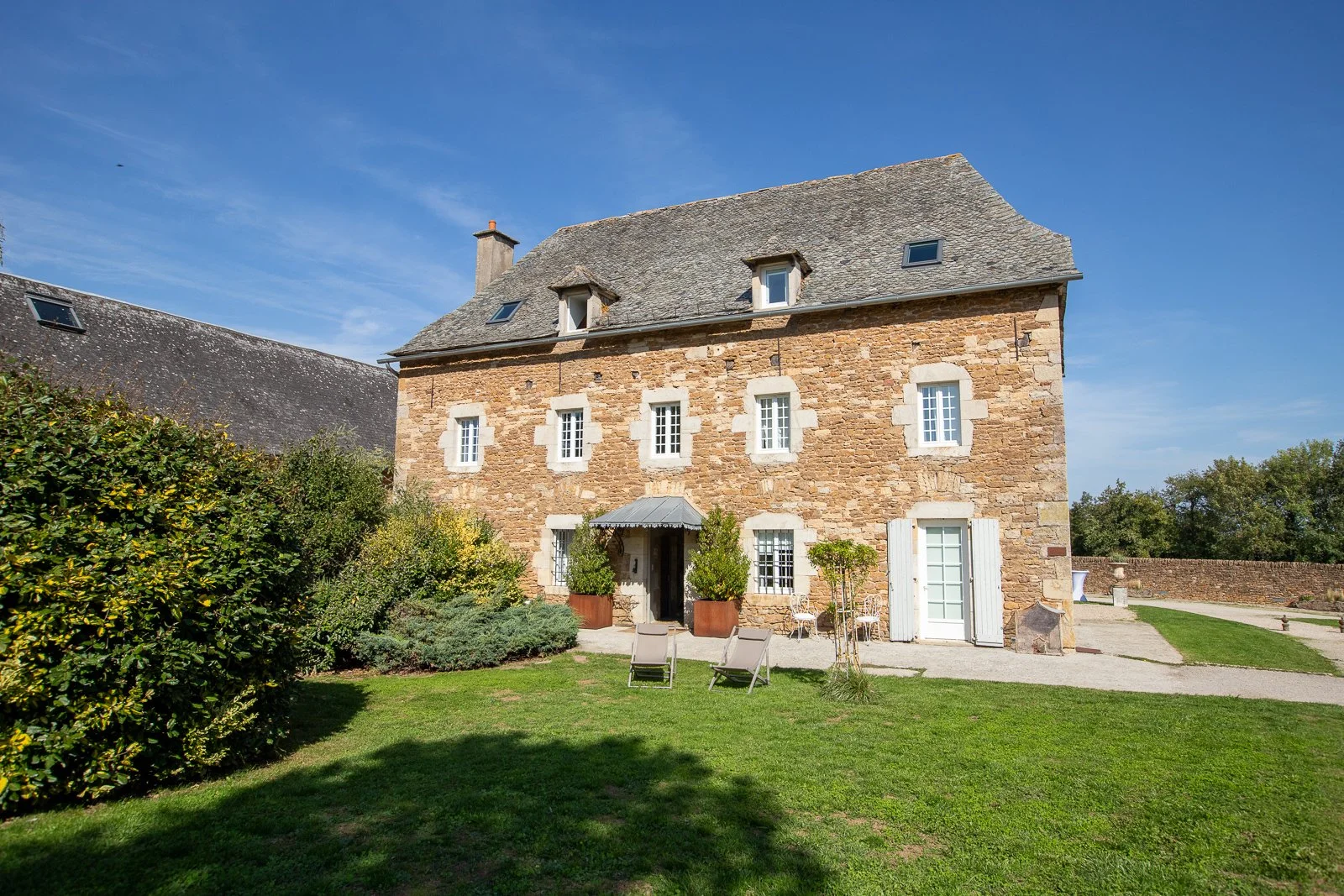 Ancienne maison en pierre avec un toit en ardoise, fenêtres à volets blancs et un jardin avec chaises longues, arbres et buissons, sous un ciel bleu.