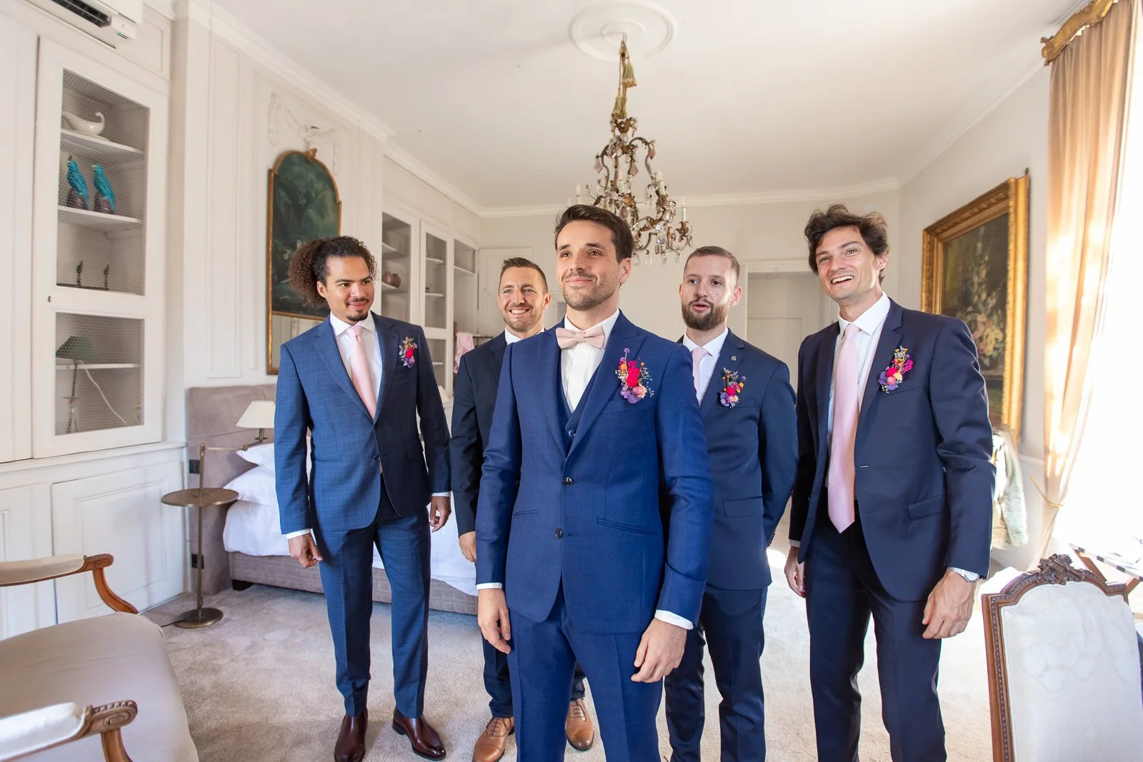 Groupe de six hommes en costumes formels, probablement lors d'un mariage, dans une pièce élégante avec lustre et peinture dans le fond.