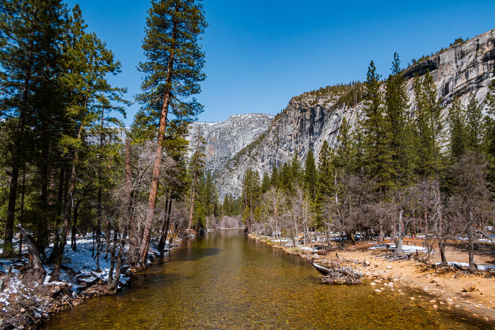 Vue d'une rivière bordée d'arbres vers une montagne rocheuse en arrière-plan, avec un ciel bleu clair, en hiver ou début du printemps.