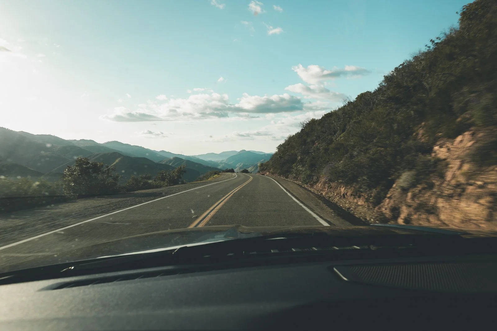 La vue depuis l’intérieur d’une voiture montrant une route sinueuse traversant des montagnes, avec des collines verdoyantes et un ciel partiellement nuageux.